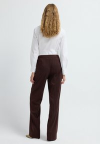 Vrouw met krullend blond haar, gekleed in een witte shirt met lange mouwen en een bruine streepbroek, staand met haar gezicht wegdraaiend tegen een witte achtergrond.