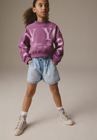 Felpa viola con testo "NY" e "Ative Stud", abbinata a pantaloni shorts di jeans chiari. Presenta sneakers alte a fantasia e calzini bianchi.