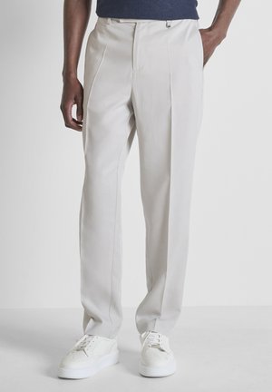Man die lichtbeige op maat gemaakte broek en witte sneakers draagt, staat met één hand in de zak tegen een effen achtergrond.