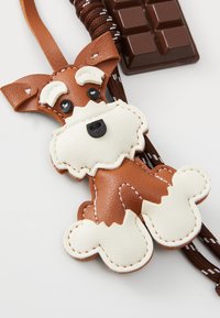 Charm en cuir en forme de chien avec des couleurs marron et blanc, présentant des détails cousus, des yeux en boutons noirs et un cordon marron.