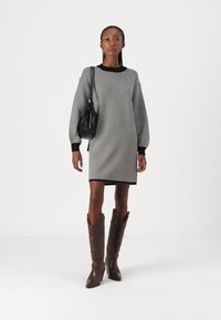 Vestido em padrão de espinha de peixe preto e branco, com mangas longas, punhos canelados, combinado com botas de cowboy castanhas e uma mala preta de ombro.