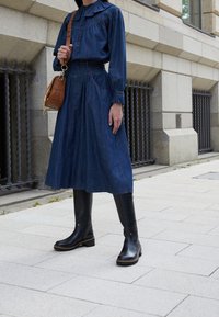 Abito in denim con maniche lunghe, colletto a volant, vita stretta e gonna a lunghezza caviglia; abbinato a stivali neri sopra il ginocchio e una borsa a tracolla marrone.
