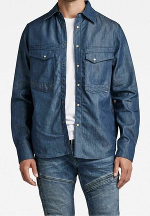Homme portant une chemise en denim bleu foncé à boutons avec poches à rabat sur la poitrine, par-dessus un t-shirt blanc, assortie à un jean en denim bleu avec des détails zippés.