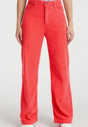 Jasnoczerwone sztruksowe spodnie noszone przez osobę stojącą z rękami swobodnie opuszczonymi po bokach, pokazane od talii do stóp.