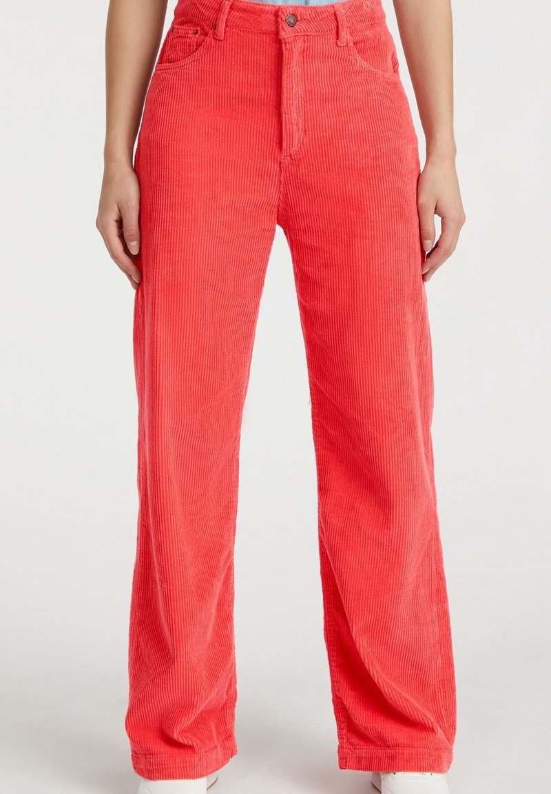 Pantalon en velours côtelé rouge vif porté par une personne debout, les mains détendues le long du corps, visible de la taille aux pieds.