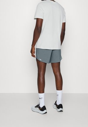 Eine Person steht mit dem Rücken zur Kamera und trägt ein weißes T-Shirt, graue Sportshorts, weiße Nike-Socken und schwarz-weiße Laufschuhe.