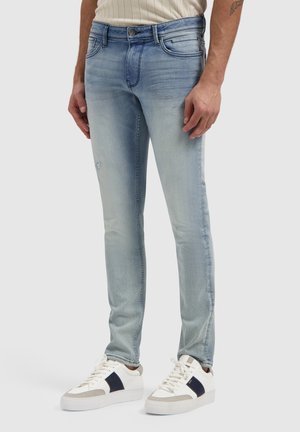 Man wearing light blue slim-fit jeans with slight distressing, white sneakers with black and beige details, standing on gray background.