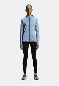 Ljusblå zip-up jacka med huva, svarta detaljer och långa ärmar. Kombinerad med svarta leggings och gråa träningsskor.