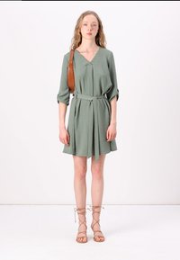 Vestido verde, até aos joelhos, com decote em V e mangas três quartos, atado à cintura. Usado com sandálias de pele claras com atacadores. Tecido simples e texturizado.