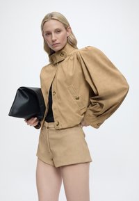 Ξανθιά γυναίκα που φοράει μπεζ τζάκετ με φουσκωτά μανίκια και ταιριαστά σορτς, κρατώντας μια μεγάλη μαύρη τσάντα φάκελο, μπροστά σε απλό λευκό φόντο.