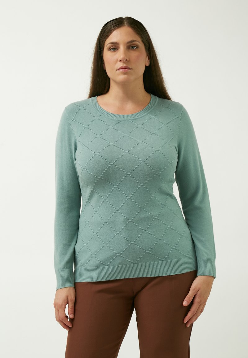 Sweater i blød teal med ribbet halsudskæring og ærmer. Har et geometrisk mønster med diamantformer. Sammen med brune bukser.