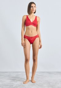 Rotes Dessous-Set mit einem glatten BH mit gewellten Kanten und Spitze. Der passende Tanga zeigt florale Spitzenmuster und ein Schleifenakzent.