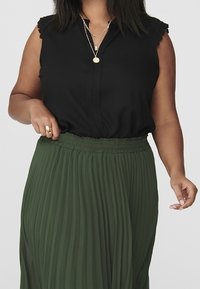 Kvinna som bär en ärmlös svart blus med spetsdetaljer och lager av guldhalsband, matchad med en grön plisserad kjol med elastisk midja.