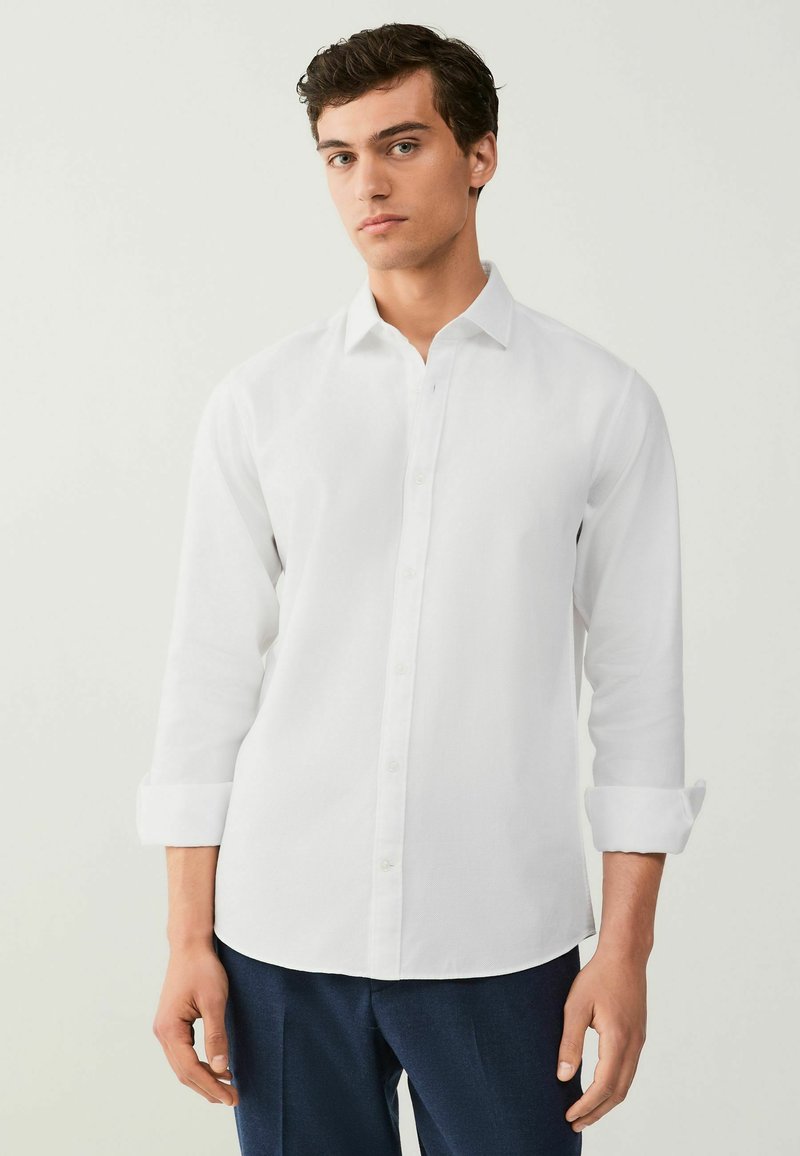 Chemise blanche à manches longues et boutons, fabriquée à partir d'un tissu lisse, dotée d'un col classique, de poignets retroussés et d'un ourlet arrondi.