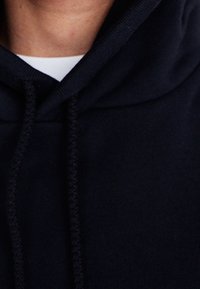 Felpa blu navy con cappuccio, dotata di cordoncino, polsini a coste e un tessuto morbido e texture. Primo piano dei dettagli del colletto e del cappuccio.