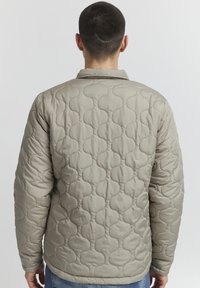 Veste beige matelassée avec un motif texturé en forme de bulles, ourlet arrondi et col classique, vue de dos.