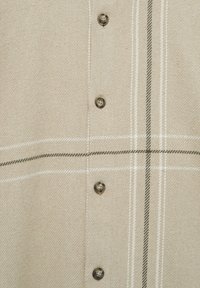 Chemise à carreaux beige avec une finition texturée, présentant des lignes vert foncé et blanches. Six boutons ronds marron sont alignés au centre.