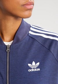 Námořnická zipová bunda s texturovaným povrchem, s třemi bílými pruhy na rukávech a bílým logem Adidas na hrudi.