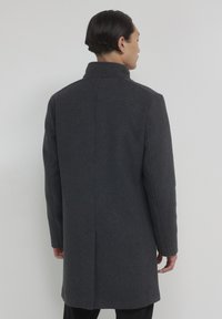 Uomo che indossa un cappotto grigio scuro lungo fino al ginocchio, in piedi contro uno sfondo grigio chiaro uniforme, visto da dietro.