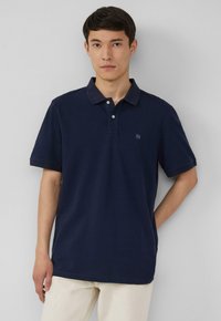 Polo-shirt à manches courtes bleu marine en coton, avec un col classique et un petit logo sur la poitrine gauche. Texture légère, coupe décontractée.