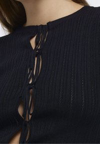 Svart strukturerad topp med vertikala ribbor, rund halsringning och en framknäppning med band som skapar en nyckelhålsöppning.