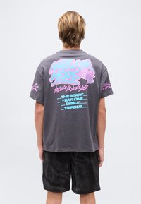 Άτομο με κοντά ξανθά μαλλιά που φοράει γκρι μπλουζάκι με ροζ και μπλε γραφικά αστεριού και κείμενο, συνδυασμένο με μαύρη υφασμάτινη βερμούδα.