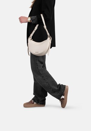 Borsa a tracolla in pelle crema con design curvo, tracolla regolabile e un logo discreto. Indossata con jeans larghi neri e sneakers marroni.