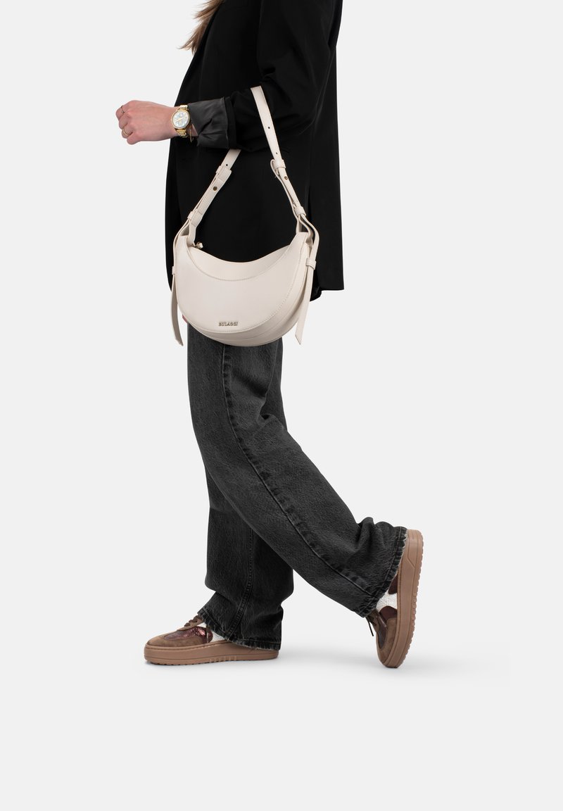 Borsa a tracolla in pelle crema con design curvo, tracolla regolabile e un logo discreto. Indossata con jeans larghi neri e sneakers marroni.