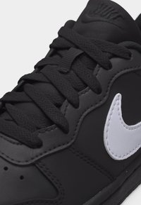 Μαύρο αθλητικό παπούτσι Nike με λευκό λογότυπο swoosh, μαύρα κορδόνια και επενδυμένη γλώσσα με ανάγλυφο λογότυπο, λεπτομερής κοντινή λήψη.