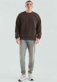 Mężczyzna stoi ubrany w brązowy sweatshirt, wyblakłe niebieskie dżinsy i białe trampki na tle jasnego, jednolitego tła.