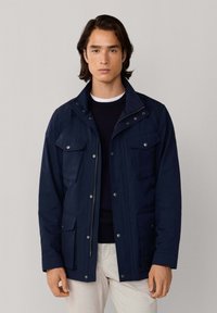 Chaqueta azul marino con cierre frontal de cremallera, broches a presión y cuatro bolsillos delanteros. Cuenta con un cuello estructurado y mangas largas, fabricada con material resistente.