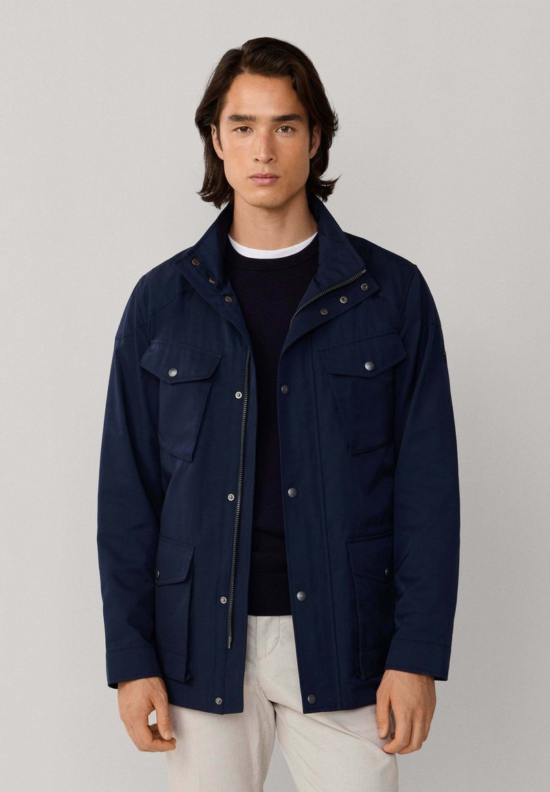 Chaqueta azul marino con cierre frontal de cremallera, broches a presión y cuatro bolsillos delanteros. Cuenta con un cuello estructurado y mangas largas, fabricada con material resistente.