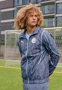 Giacca Puma con un motivo mimetico grigio e accenti blu, con zip frontale e logo. Tessuto morbido con un design sportivo adatto all'uso atletico.