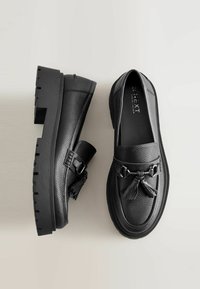 Mocasines de cuero negro con un acabado texturizado, suela de plataforma gruesa, borlas decorativas y detalle en metal plateado.