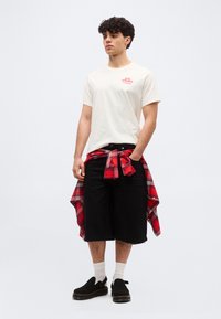 Νεαρός άνδρας φοράει λευκό μπλουζάκι Levi's, μαύρα σορτς μέχρι το γόνατο, μαύρα παπούτσια τύπου slip-on, λευκές κάλτσες και κόκκινο καρό πουκάμισο δεμένο στη μέση.