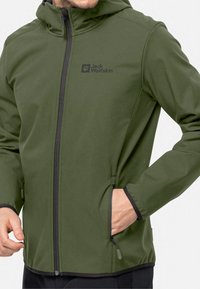Veste softshell vert olive avec une fermeture éclair avant, une capuche et deux poches latérales. Accents noirs le long de la fermeture éclair et des poignets. Logo Jack Wolfskin affiché.