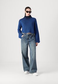 Blå rillede turtleneck sweater med brede ærmer, parret med højtaljede lyse denim jeans og hvide platform sneakers. Sorte solbriller inkluderet.