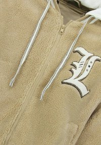 Beige fleecejack met ritssluiting, voorzien van een geborduurde letter "L" en een capuchon met trekkoord, met een zachte textuur en een casual ontwerp.