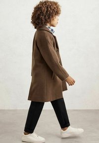 Rudas vilnonis paltas su pritaikytu dizainu, šoniniais kišenėmis, derinamas su juodomis kelnėmis ir baltais sportbačiais. Vaikas vaikšto šoniniu vaizdu.