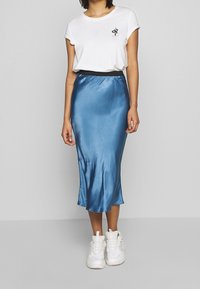 Weißes Baumwoll-T-Shirt mit kleinem schwarzem Motiv, kombiniert mit einem knieumspielenden, glänzenden blauen Satinhrock und weißen Turnschuhen.