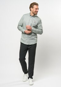 Camicia button-up verde chiaro con un piccolo logo, abbinata a pantaloni grigio scuro e sneakers bianche, in piedi contro uno sfondo neutro.