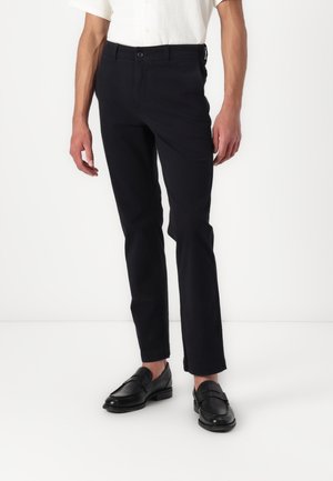 Mann trägt schwarze Slim-Fit-Hose, weißes Hemd in die Hose gesteckt und schwarze Lederschuhe, steht vor einem schlichten weißen Hintergrund.