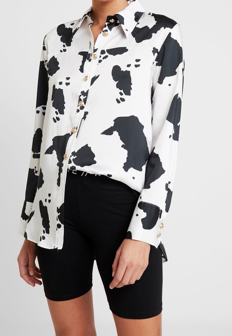 Femme portant une chemise blanche avec de grandes taches noires à imprimé vache et un short noir, debout devant un fond uni clair.