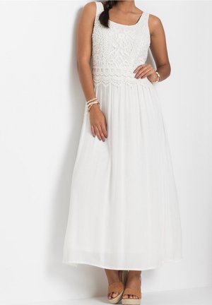 Femme portant une robe maxi blanche sans manches avec un corsage en dentelle et une jupe fluide, accessoirisée avec des bracelets et des sandales compensées marron.