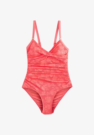 Femme aux cheveux bouclés portant un maillot de bain une pièce corail texturé, debout contre un mur blanc, les mains dans le dos.