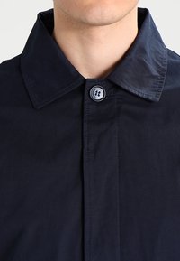 Marineblå bomuldsskjorte med spids krave, der har lukning med knapper og en diskret tekstur. Stoffet virker glat og let.