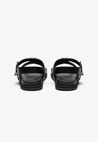 Svarte skinnsandaler med justerbare spenner, flate såler og minimalistisk design. Har en glatt tekstur og åpen bak for pusteevne.