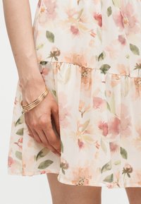 Χέρι που φοράει χρυσά βραχιόλια ακουμπάει δίπλα σε ανοιχτό φόρεμα με λουλουδένιο σχέδιο σε ροζ, ροδακινί και πράσινα χρώματα.