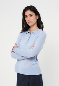 Ανοιχτό μπλε, μακρυμάνικο πόλο πουκάμισο με κολάρο με κουμπιά, μαλακή υφή και ριμπ τελείωμα, φορεμένο με σκούρα κάτω ρούχα.