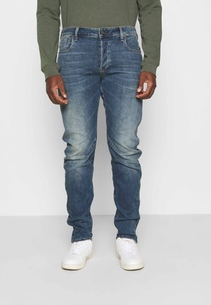 Mężczyzna w wyblakłych niebieskich dżinsach, oliwkowozielonej koszuli z długim rękawem i białych tenisówkach, stoi na tle jednolitego, jasnego tła.